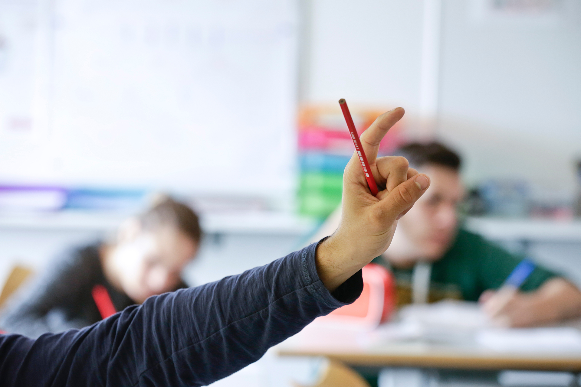Hand mit nach oben ausgestrecktem Zeigefinger und Bleistift in einem Klassenzimmer