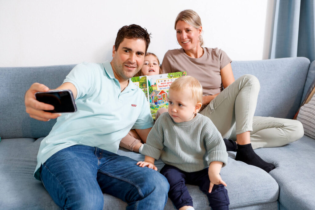 Ein blinder Mann macht mit seiner Frau und seinen zwei kleinen Kindern im Wohnzimmer am Sofa ein Selfie.